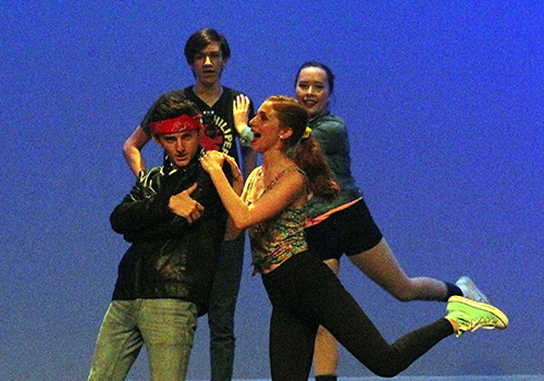 Sophomores Cry Lester, Sydney Rostedt, Chase Carson, and Mckenna Bonney lean back and forward on stage under the spotlight.
