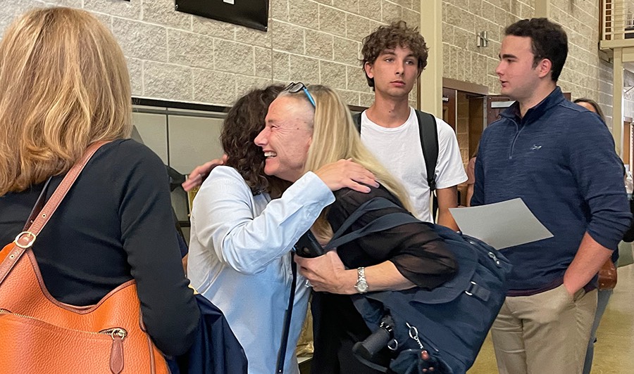 The ceremony is moved outside the auditorium for family and friends to socialize and take pictures when two women recognize each other and embrace the friendly face with a hug. 